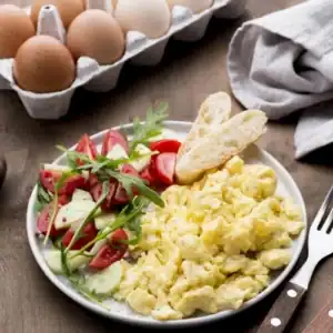 A plate of scrambled eggs served with a fresh tomato, cucumber, and arugula salad, alongside two slices of crusty bread. A carton of eggs is in the background.
