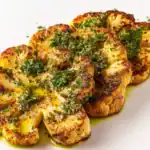 A close-up view of perfectly roasted cauliflower steaks garnished with fresh herbs and drizzled with a vibrant green sauce on a white plate.