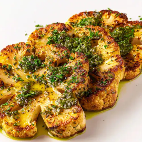 A close-up view of perfectly roasted cauliflower steaks garnished with fresh herbs and drizzled with a vibrant green sauce on a white plate.
