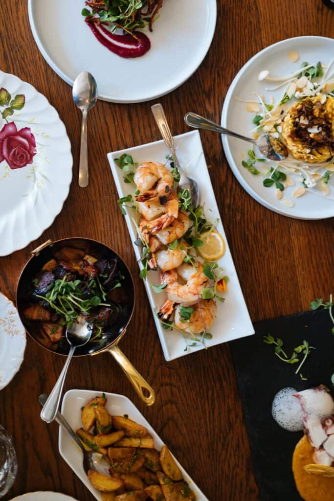 An elegant spread of dishes on a wooden table, featuring lemon herb grilled shrimp garnished with microgreens, roasted vegetables, and other gourmet accompaniments.