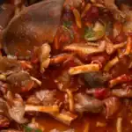 Close-up of a rich mushroom and tomato-based sauce with chunks of mushrooms, herbs, and a wooden spoon.