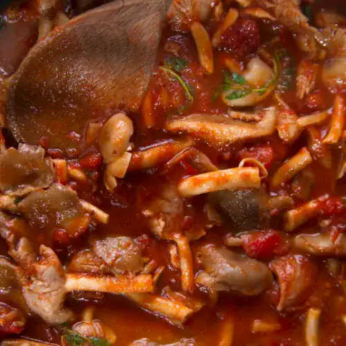 Close-up of a rich mushroom and tomato-based sauce with chunks of mushrooms, herbs, and a wooden spoon.
