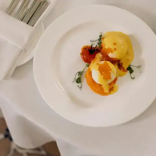 A plated dish featuring poached eggs topped with hollandaise sauce and orange caviar, served with lemon brioche and garnished with greens.