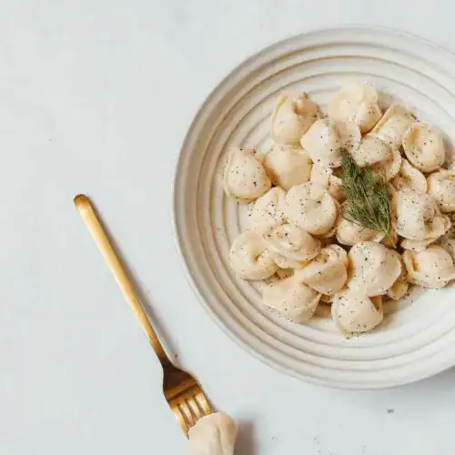 A white plate filled with creamy pumpkin ravioli garnished with fresh dill and a sprinkle of black pepper, accompanied by a gold fork on a marble surface.
