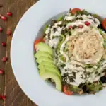 A colorful quinoa and kale salad topped with sliced avocado, pomegranate seeds, and a creamy dressing, served in a white plate.