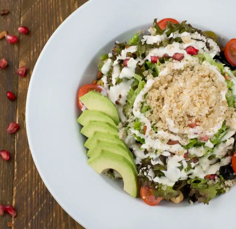 A colorful quinoa and kale salad topped with sliced avocado, pomegranate seeds, and a creamy dressing, served in a white plate.