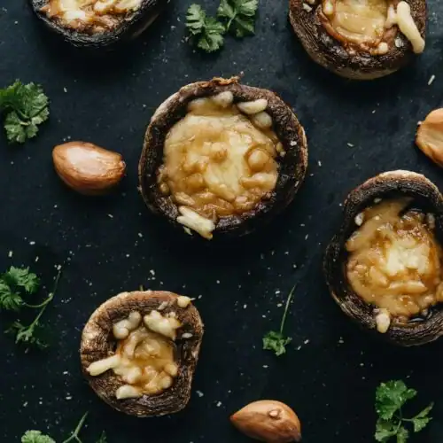 Baked Portobello mushrooms filled with goat cheese and garnished with fresh parsley and roasted garlic cloves, arranged on a black slate surface.