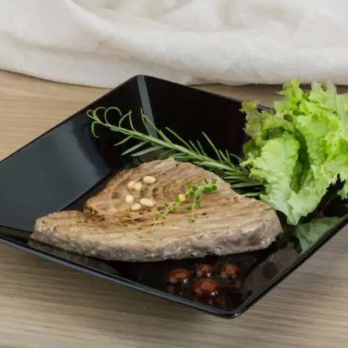 A perfectly seared sesame-crusted tuna steak served on a black square plate, garnished with fresh herbs and accompanied by crisp lettuce.