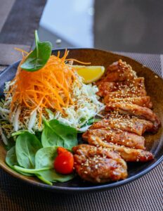 A plate of swicy Korean chicken garnished with sesame seeds, served with fresh cabbage salad, shredded carrots, spinach leaves, and a lemon wedge on a dark ceramic dish