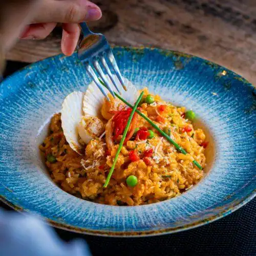 A vibrant plate of saffron risotto topped with pan-seared scallops, garnished with peas, red peppers, Parmesan, and fresh chives, served in an elegant blue ceramic dish