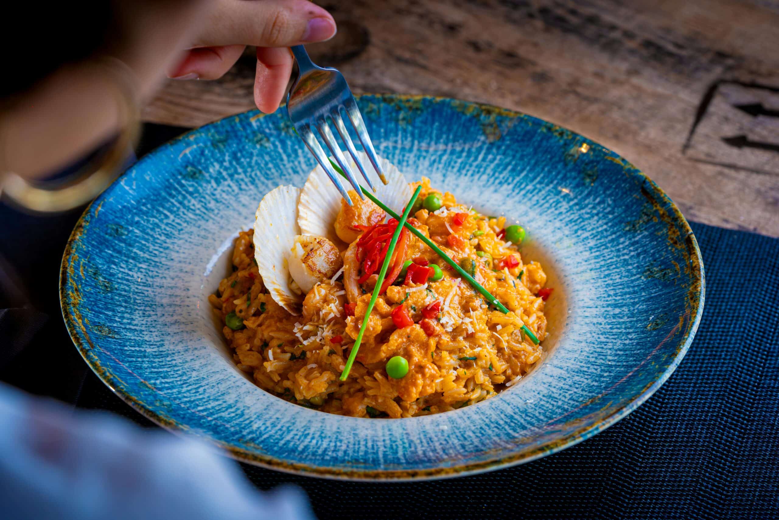 A close-up of pan-seared scallops served over saffron risotto with vegetables, garnished with chives in a blue ceramic plate.