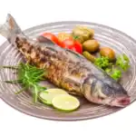 A rbaked whole sea bass on a gray plate, surrounded by garnishes of fresh rosemary, lime slices, cherry tomatoes, olives, parsley, and a pickle.