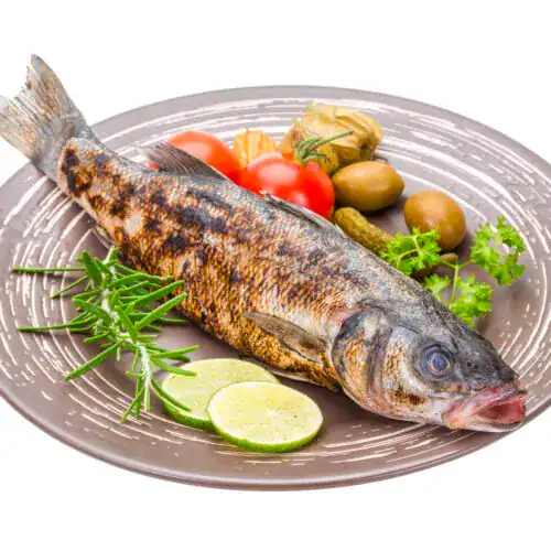 A rbaked whole sea bass on a gray plate, surrounded by garnishes of fresh rosemary, lime slices, cherry tomatoes, olives, parsley, and a pickle.