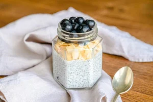 A jar of creamy chia pudding topped with toasted coconut flakes and fresh blueberries, placed on a wooden table with a spoon and a light cloth.