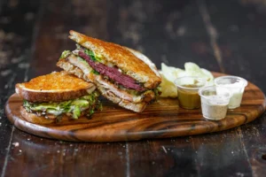 A gourmet sandwich with beef pastrami, lettuce, and cheese served on a wooden board with a side of dipping sauces and chips.