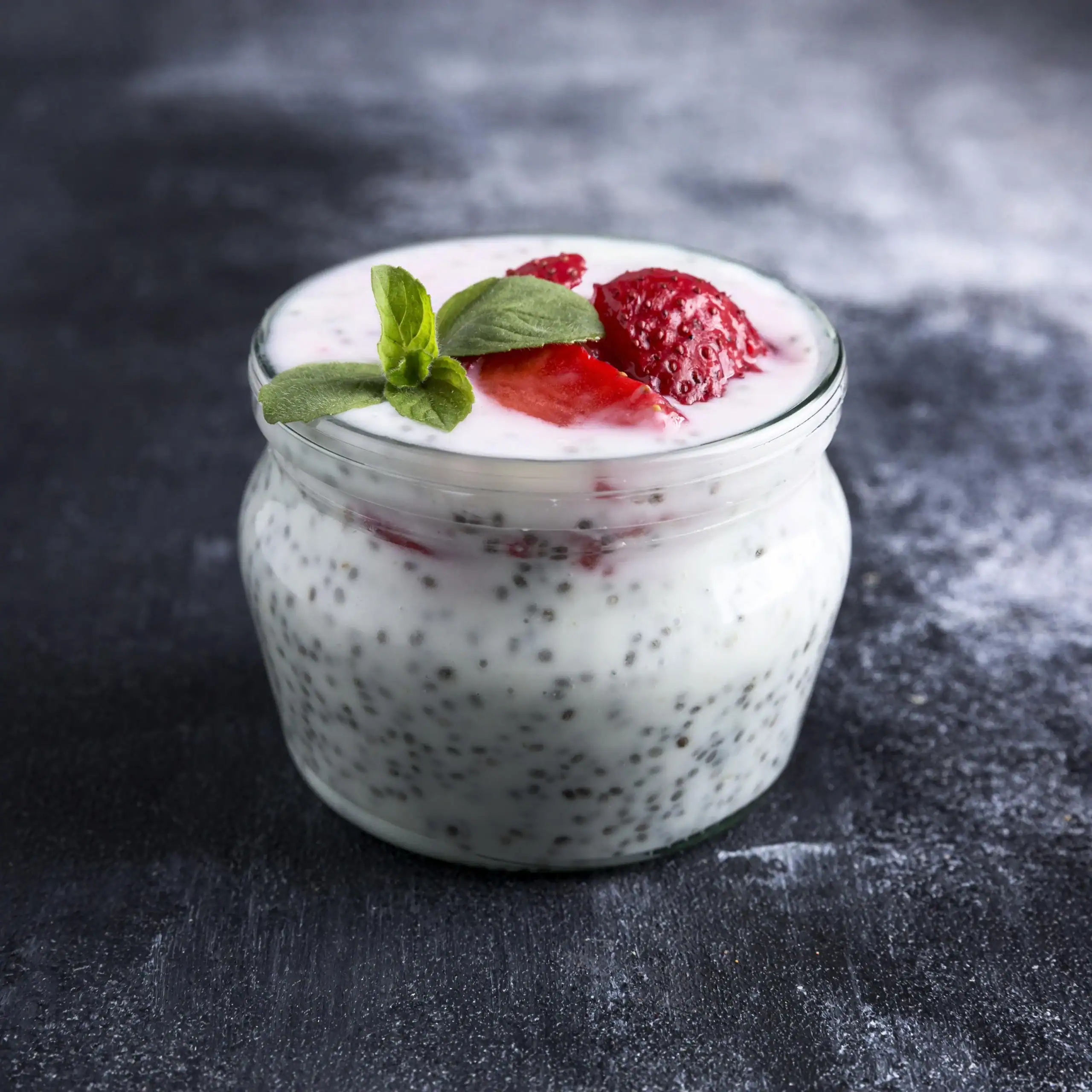 A jar of creamy chia pudding topped with fresh strawberries and a mint sprig, placed on a dark textured surface.