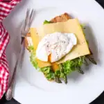 A beautifully plated toast topped with grilled asparagus, fresh lettuce, a slice of cheese, and a perfectly poached egg, served on a white plate with a red checkered napkin.