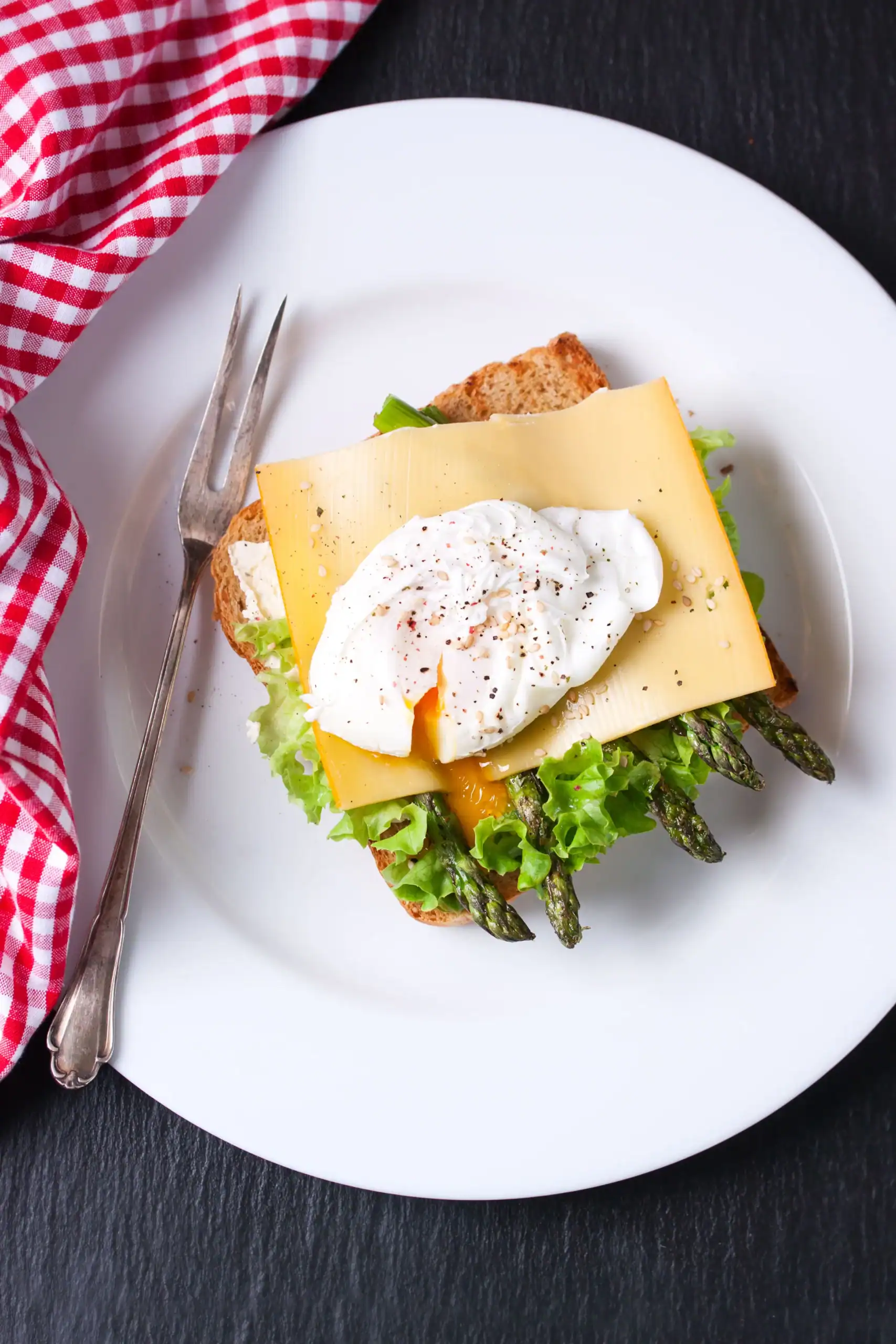 A beautifully plated toast topped with grilled asparagus, fresh lettuce, a slice of cheese, and a perfectly poached egg, served on a white plate with a red checkered napkin.
