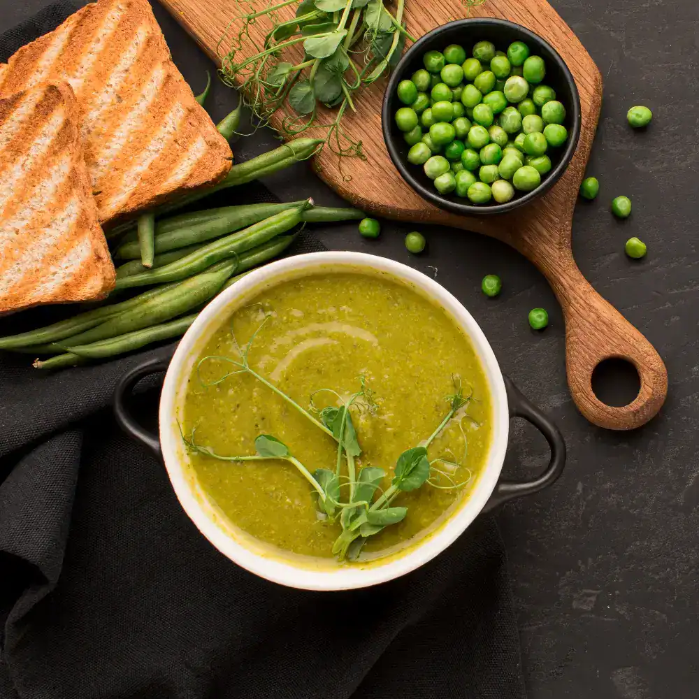 A bowl of creamy split pea soup garnished with fresh pea shoots, served with toasted bread slices, fresh peas, and green beans on a wooden cutting board over a dark background.