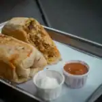 A sliced chicken Mexican breakfast burrito served on a metal tray, accompanied by sour cream and salsa in small cups.