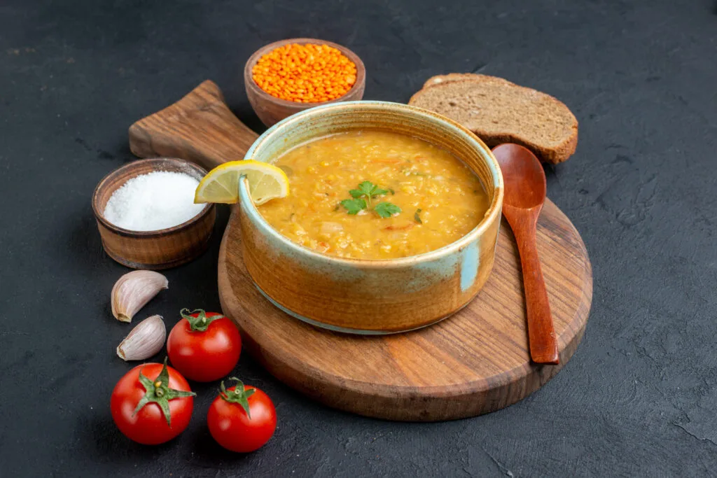 A bowl of classic lentil soup garnished with fresh parsley and lemon, served with bread, tomatoes, and garlic on a wooden board.