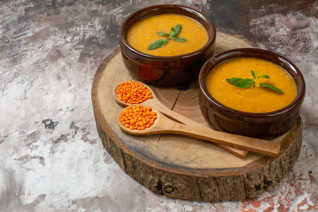 Two bowls of lentil soup garnished with fresh basil leaves, served on a rustic wooden slab with spoons of red lentils nearby.