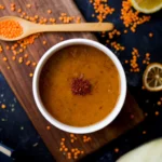 A bowl of lentil soup topped with chili flakes, surrounded by red lentils, lemon slices, and a wooden spoon on a rustic wooden board.