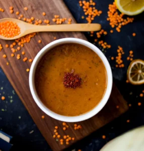 A bowl of lentil soup topped with chili flakes, surrounded by red lentils, lemon slices, and a wooden spoon on a rustic wooden board.