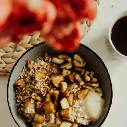A top view of a bowl of oatmeal topped with caramelized apple chunks, cashews, a dollop of cream, and a sprinkle of nuts, alongside a cup of tea and blurred flowers in the foreground.