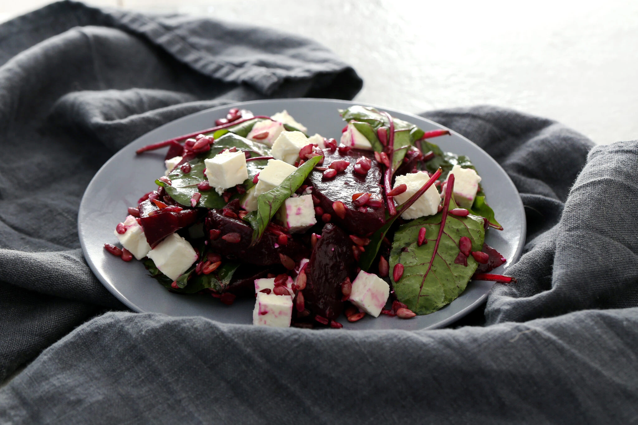 A plate of beetroot salad with fresh greens, feta cheese, and pomegranate seeds, served on a gray plate with a dark fabric backdrop.