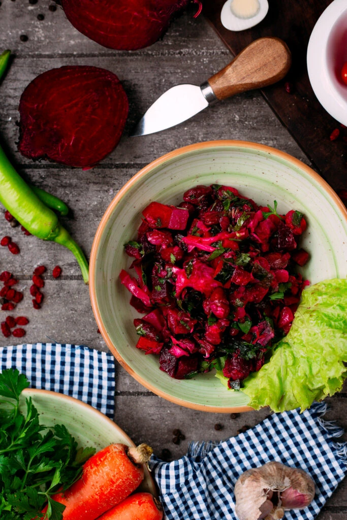 A vibrant bowl of beetroot salad garnished with fresh herbs and lettuce, surrounded by raw vegetables like carrots, garlic, and sliced beets on a rustic table.