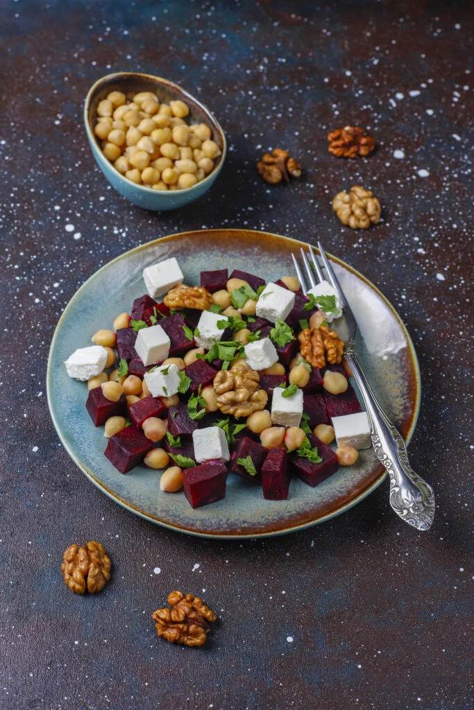 A plate of beet salad with diced beets, chickpeas, feta cheese, walnuts, and fresh herbs, served with a fork on a textured dark surface, with additional walnuts and chickpeas as garnish.