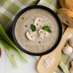 A bowl of creamy mushroom soup garnished with sliced mushrooms and fresh parsley, accompanied by fresh bread, green onions, and whole mushrooms on a wooden table.