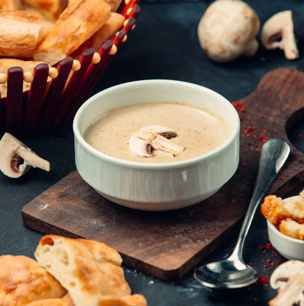 A bowl of creamy mushroom soup garnished with a single slice of mushroom, served on a wooden board with fresh bread, croutons, and mushrooms in the background.