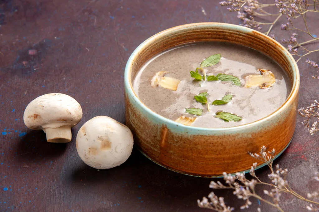 A bowl of rich, dark mushroom soup garnished with fresh mint leaves and mushroom slices, surrounded by whole mushrooms and dried flowers on a rustic background.