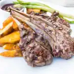 Juicy grilled lamb chops seasoned with rosemary, served alongside crispy potato wedges, tender asparagus, and a side of sauce on a white plate.