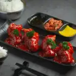 A black rectangular plate holding Korean fried chicken wings glazed with a vibrant sweet and spicy sauce, garnished with parsley, served alongside traditional Korean side dishes.