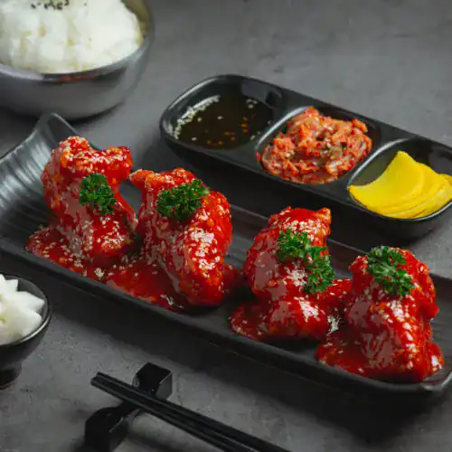 A black rectangular plate holding Korean fried chicken wings glazed with a vibrant sweet and spicy sauce, garnished with parsley, served alongside traditional Korean side dishes.