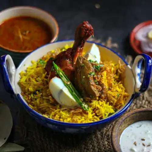 A bowl of lamb biryani topped with tender lamb pieces, a drumstick, and boiled egg halves, garnished with green chilies and fresh herbs, accompanied by curry and raita. Title: Lamb Biryani Recipe | A Fragrant and Flavorful One-Pot Dish