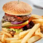 A gourmet lamb burger topped with fresh vegetables, including pickles, red onions, and lettuce, served with a side of golden French fries.