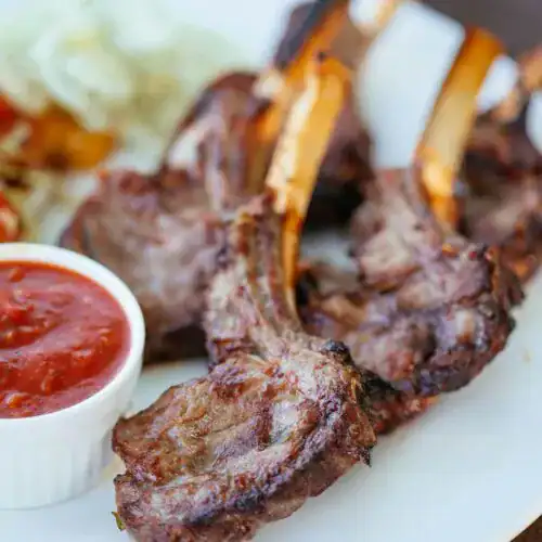 Perfectly seared lamb chops with crispy edges, served on a white plate alongside a ramekin of tomato-based dipping sauce and a fresh side salad.