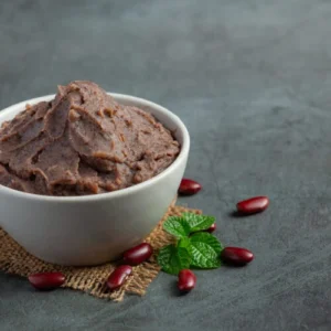 A bowl of creamy refried red beans placed on a piece of burlap, garnished with fresh mint leaves and surrounded by scattered red beans on a dark textured surface