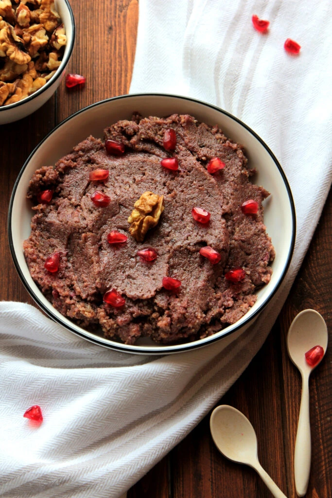 A bowl of refried beans garnished with pomegranate seeds and a walnut, placed on a wooden table with white fabric and spoons nearby.