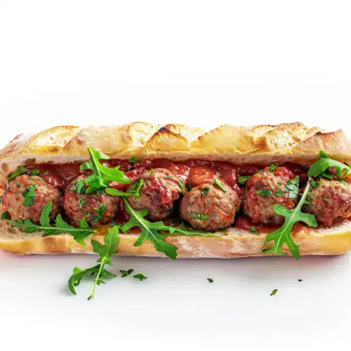 A lamb meatball sub in a crispy baguette, topped with tomato sauce, fresh arugula, and garnished with parsley.