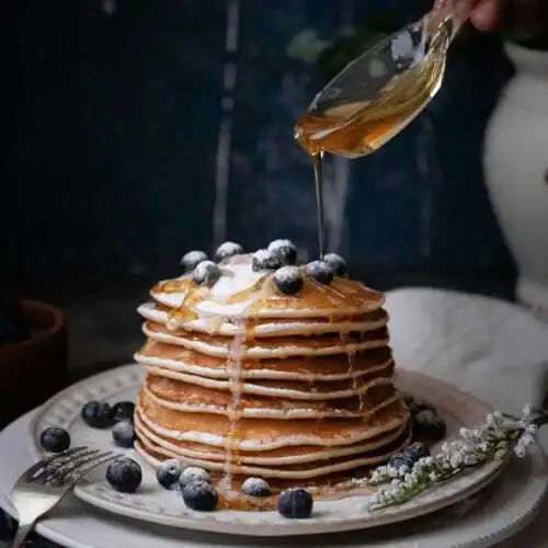 A stack of pancakes drizzled with golden raw honey, topped with fresh blueberries and a dusting of powdered sugar on a rustic plate.