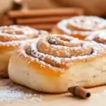 Close-up of soft Mallorca bread rolls dusted with powdered sugar, placed on a wooden board with cinnamon sticks.
