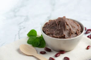 A bowl of creamy refried beans placed on a light fabric, garnished with fresh mint leaves and surrounded by red beans, with a wooden spoon nearby on a marble background.