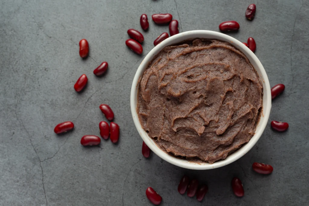 A bowl of creamy refried beans placed on a textured gray surface, surrounded by scattered red beans.