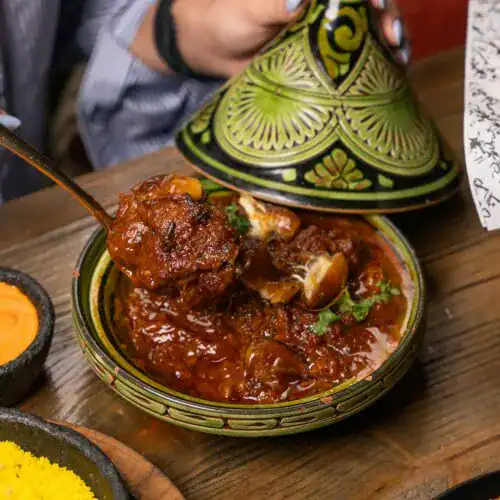 A vibrant Moroccan lamb tagine served in a traditional green and black tagine pot, with tender lamb pieces in a rich, spiced tomato-based sauce, being lifted with a spoon.