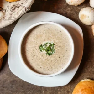 A bowl of creamy mushroom soup garnished with fresh parsley, served on a white plate and surrounded by bread rolls and whole mushrooms on a rustic table.